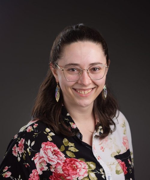 A smiling person with long dark hair, glasses, and colorful earrings wears a black and white button-up shirt with a large floral pattern against a dark, neutral background—perfect for Heller Consulting President Jeffrey Appells bio.