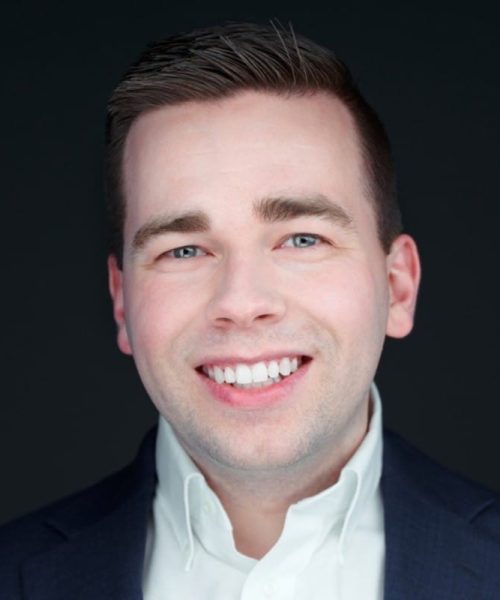 A young man with short brown hair, wearing a dark suit jacket and white collared shirt, smiles confidently in front of a plain dark background, reflecting his role as a dedicated nonprofit consultant.