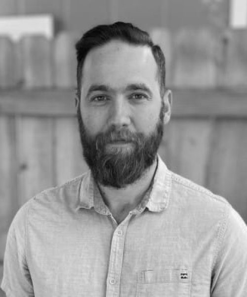 Tyler Van Kirk at Heller Consulting Expert Data stands outside in front of a wooden fence, wearing a light-colored button-up shirt. He has a beard and short hair. The image is in black and white.