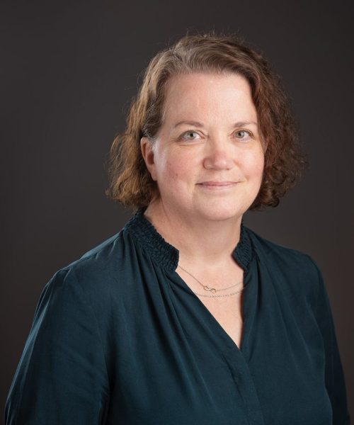 A woman with wavy brown hair wearing a dark teal blouse and a delicate necklace smiles softly while standing in front of a dark, plain background, echoing the professional poise seen in Heller Consulting President Jeffrey Appell’s bio.