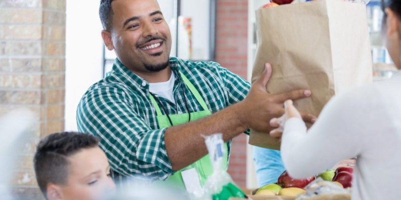 A smiling grocery store worker in a green apron hands a paper bag full of groceries to a customer. A child stands nearby at the checkout counter.