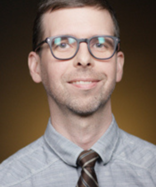 A man with short brown hair and glasses, wearing a light gray button-up shirt and a brown striped tie, smiles against a dark gradient background.