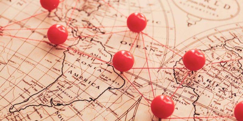 Close-up of a vintage world map with red push pins connected by red string, marking various locations across North America, Europe, and surrounding regions.