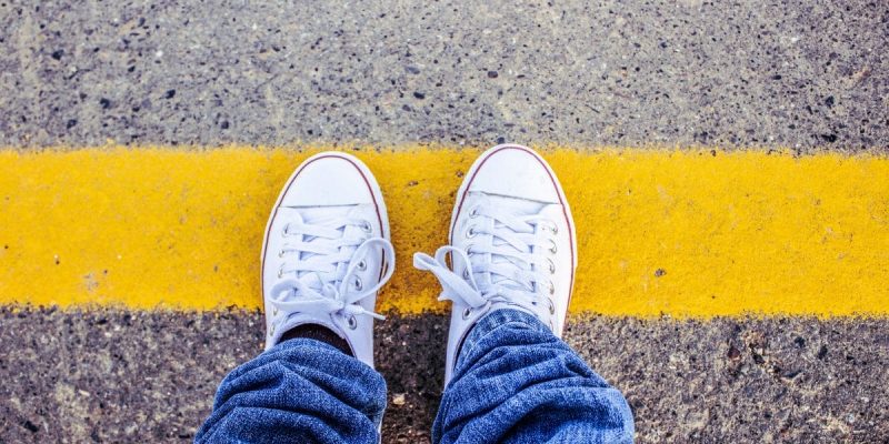 A person wearing white sneakers and blue jeans stands at a yellow line painted on a rough concrete surface, viewed from above.