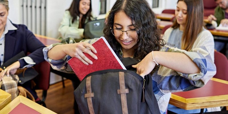 A student with curly hair and glasses smiles while placing a red notebook into her backpack in a classroom, with other students sitting at desks in the background.